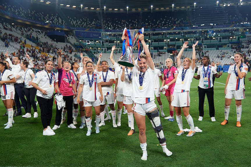 Australian footballer Ellie Carpenter lifts a cup above her head while wearing a leg brace as her teammates cheer.