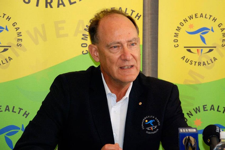 A man with a receding hairline, wearing a black blazer, sits behind microphones in front of a Commonwealth Games backdrop.
