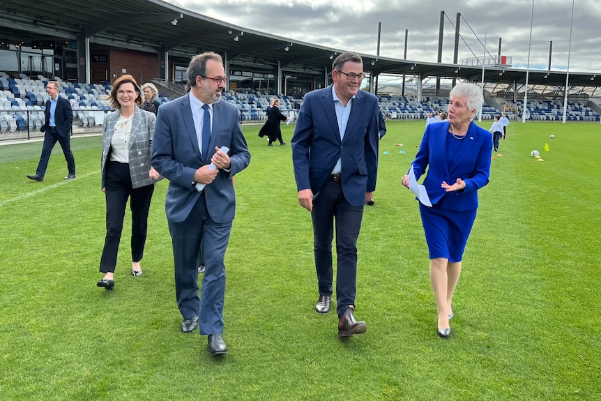 Smiling Daniel Andrews in a stadium. A man and woman in blue suits walk beside him, other people mingle behind, cloudy sky.