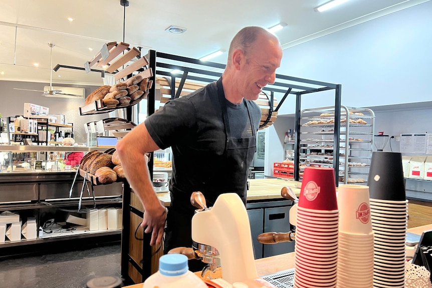 A bald man laughs as he puts on a apron in front of a coffee machine, bakery stands behind with croissants and danish.