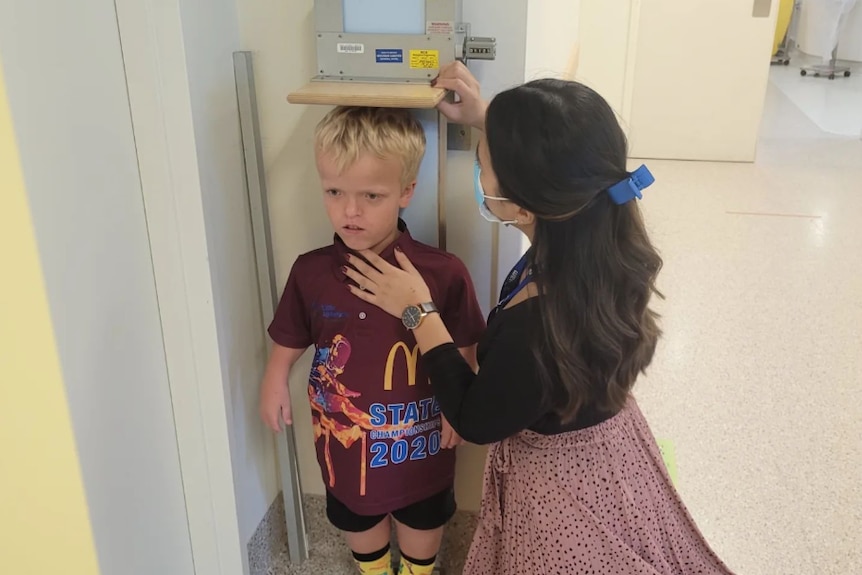 A young, fair-haired boy has his height measured by a medical professional in a clinic.