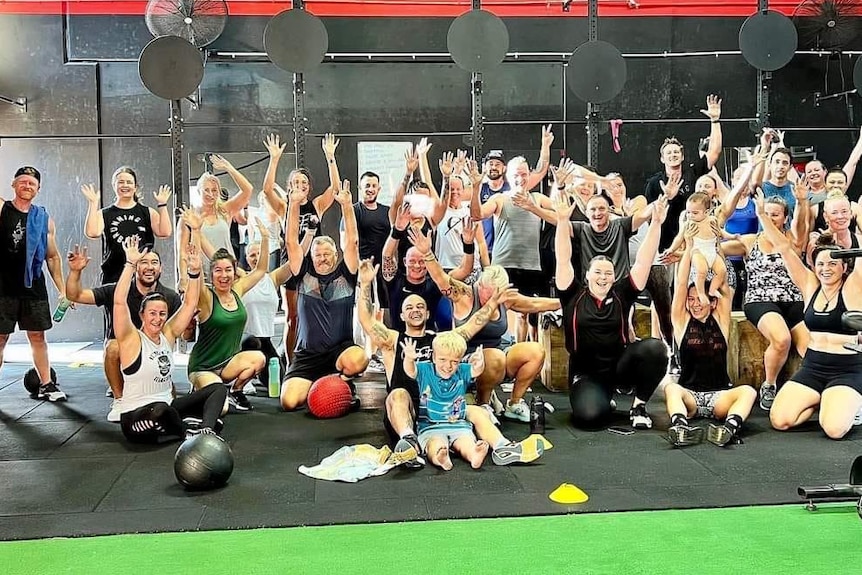 A large group of smiling people in workout gear waving their hands in a gym.