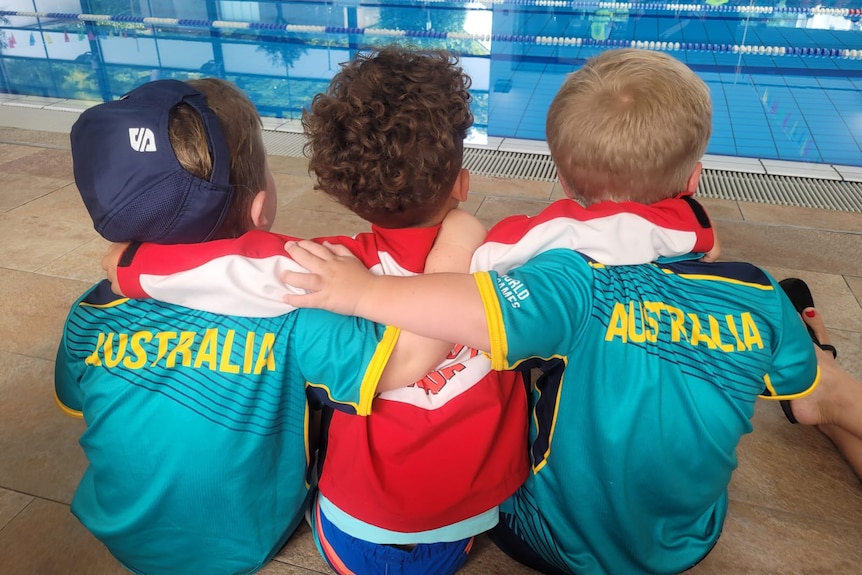 Three youngsters, two in Australian sports gear, sitting near the edge of a pool with their arms around each other.