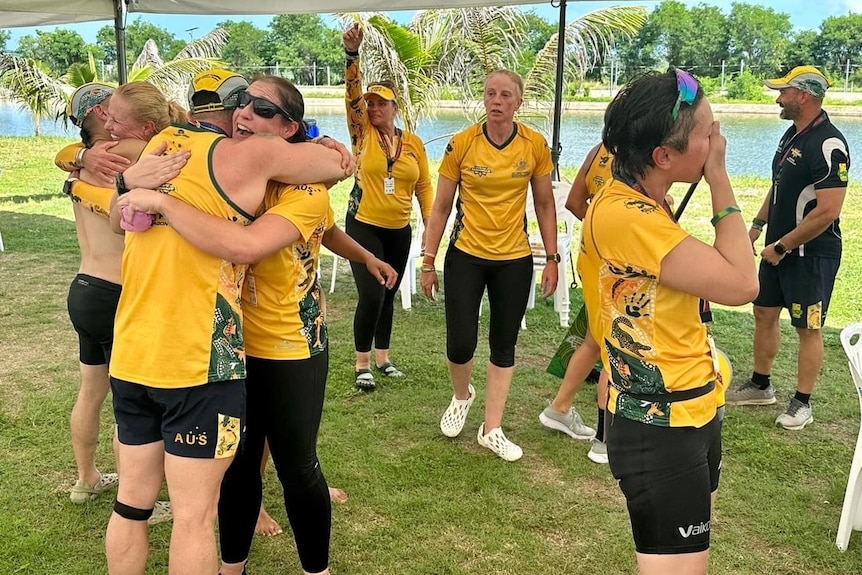 A number of women in gold jerseys standing hugging and celebrating by a river.