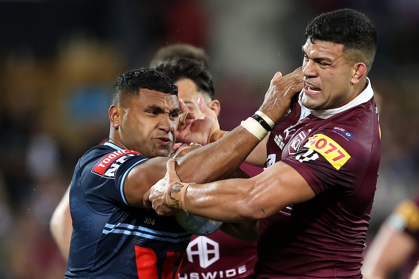 A NSW State of Origin player grimaces as he holds the ball in one hand and uses his other hand to fend off a Maroons defender.
