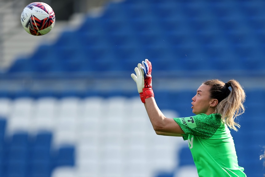 A woman goalkeeper wearing green gets ready to catch a soccer ball in a stadium