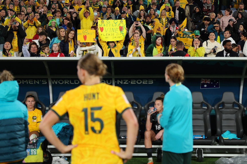 Matildas players watch on at pitchside as Australian fans in the stands cheer and hold up signs after a big match.