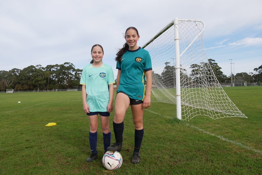 Two girls wearing Australian soccer shirts standing behind a ball. 