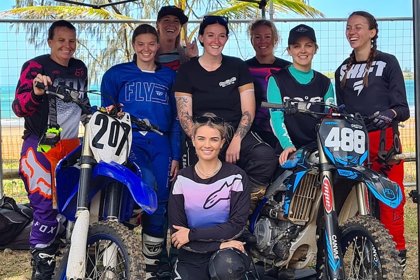 Group of women stand with two motorbikes smiling at camera. 