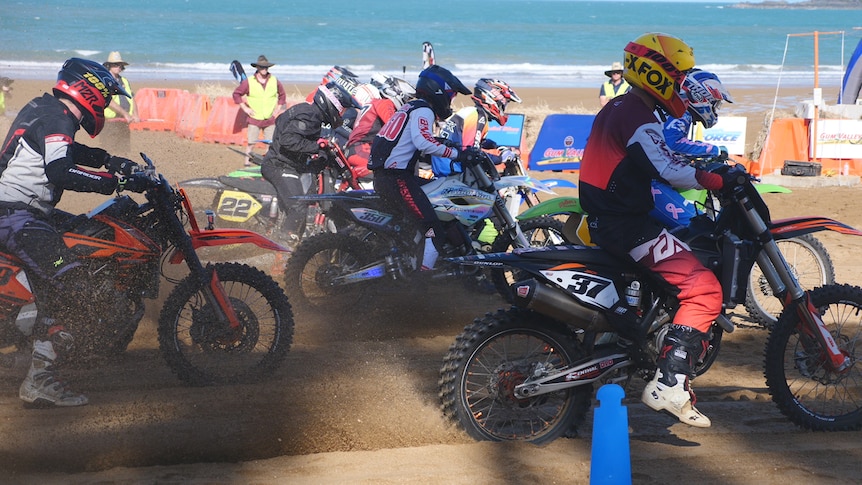 Motorbikes and riders start race side by side on the beach with sand flying behind them. 