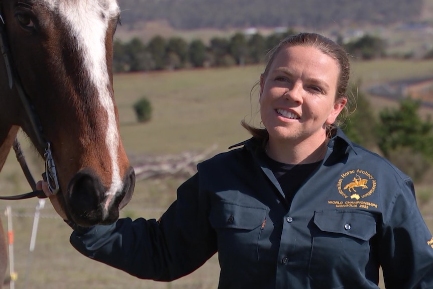 A woman with long brown hair, wearing a green collared short, smiles, as she stands beside a horse.
