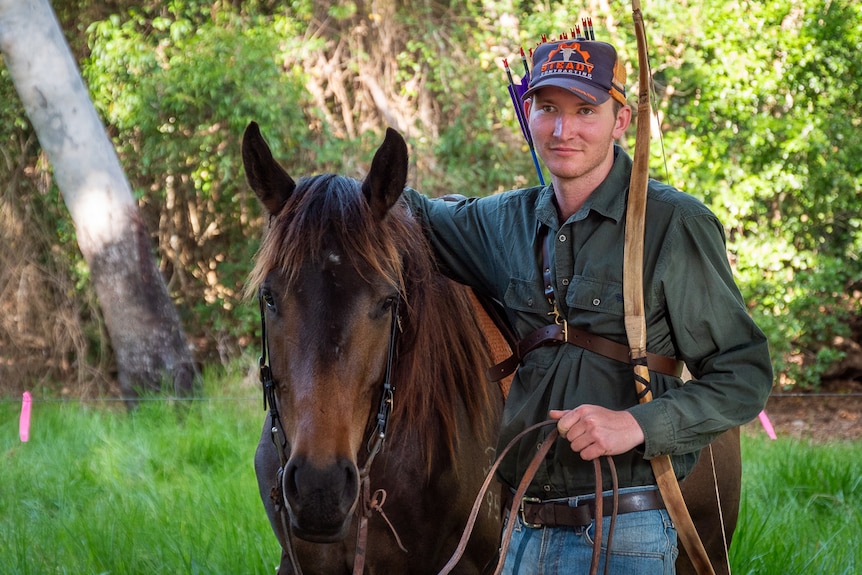 young man holding a bow with quiver of arrows on his back holding the reins of a brown horse
