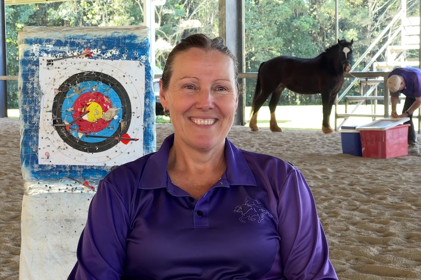 A smiling woman with brown hair, wearing a blue blouse, stands in front of a target. A horse is standing in the background.