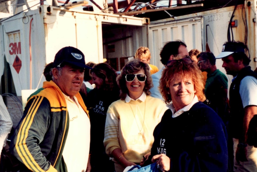A man in a tracksuit and cap – Alan Bond – stands next to a smiling blonde woman and another woman, his wife Eileen.