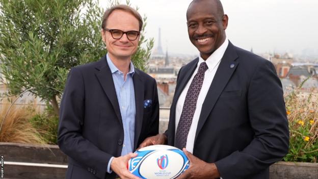 Herbert Mensah and Julien Collette stand side-by-side passing a rugby ball between them with a Paris skyline in the background