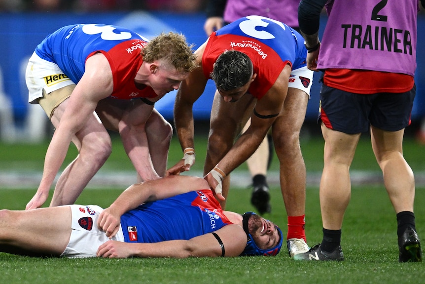 One AFL player lies motionless on the ground, two players at his aid, as a trainer runs on the ground.