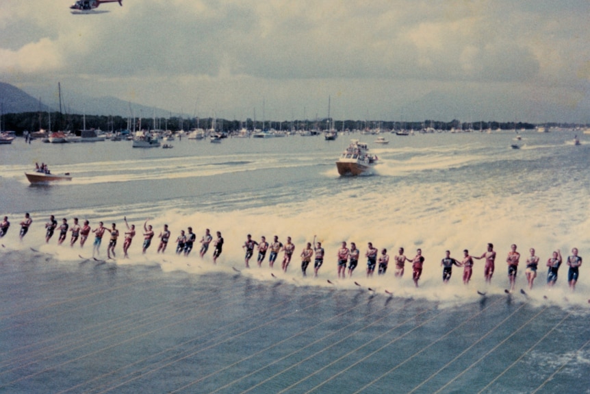 Photo of row of water skiers with boats behind and helicopter above
