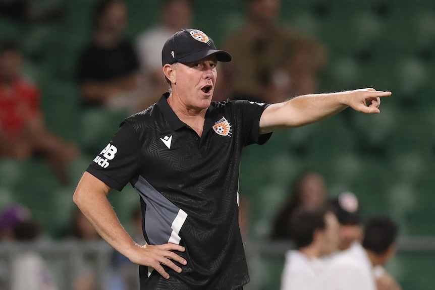 A man in a black t-shirts points from the sidelines of a sporting field. 