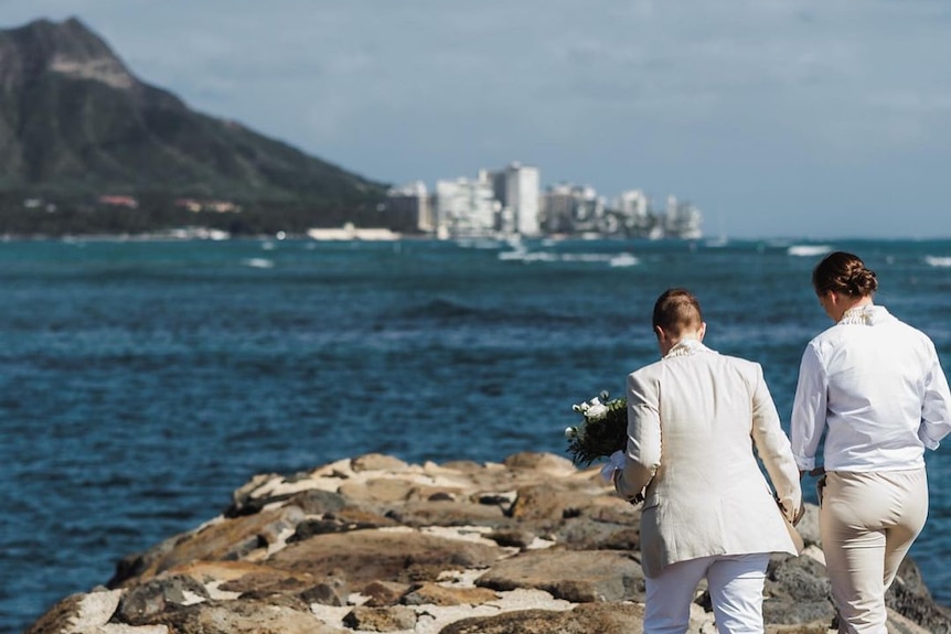 The couple walk hand in hand out onto some rocks along the water, the view of a city on another island is in the distance.