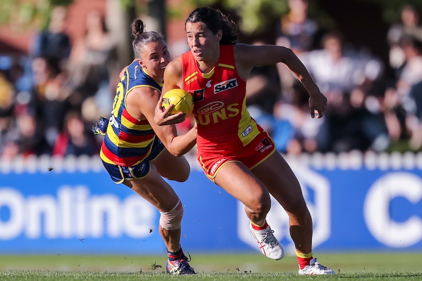 A Gold Coast Suns AFLW player holds the ball with her right hand as a Crows opponent attempts a tackle.