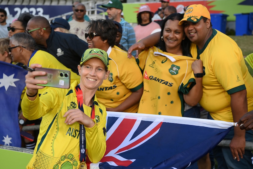 A smiling Australian skipper Meg Lanning holds a mobile phone and poses in front of a couple of Australian fans with a flag. 