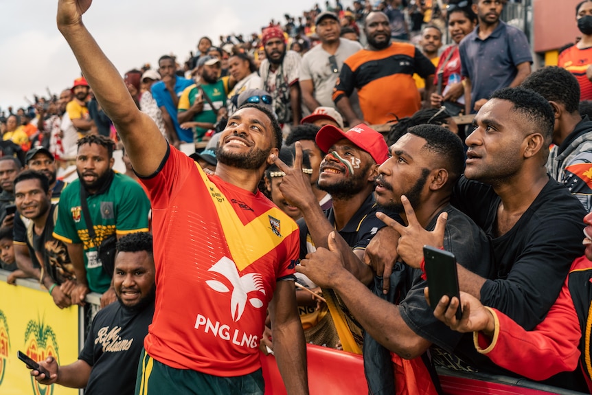 A rugby player poses for a selfie with members of the crowd, they all smile.