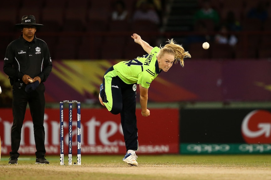 An Irish women's cricketer stares down the pitch after delivering a ball, as the umpire watches in the background.