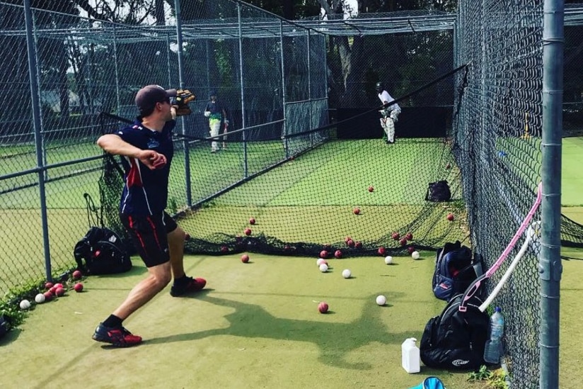 A man throwing a ball at a batsman inside cricket nets.