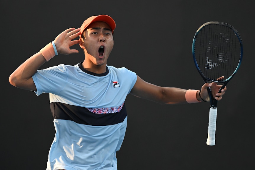An Australian male professional tennis player holds his right hand to his ear as he celebrates winning a point.