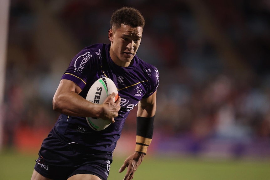 A Melbourne Storm NRL player runs the ball tucked under his right arm.