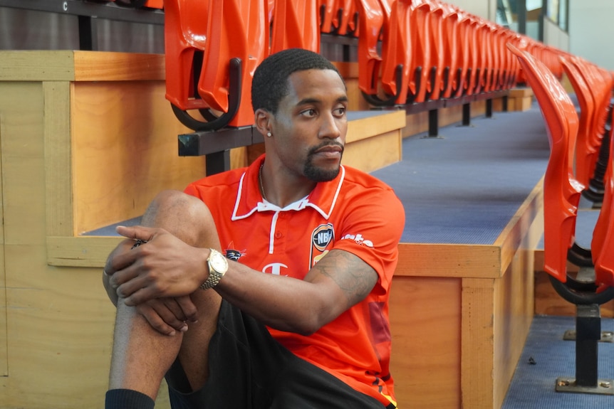 Bryce Cotton sitting on the floor next to tiered seating, and looking away from the camera.
