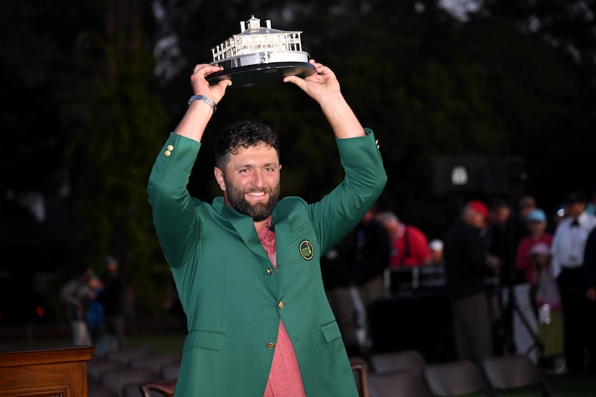 Jon Rahm smiles while holding the Masters trophy over his head, wearing the green jacket