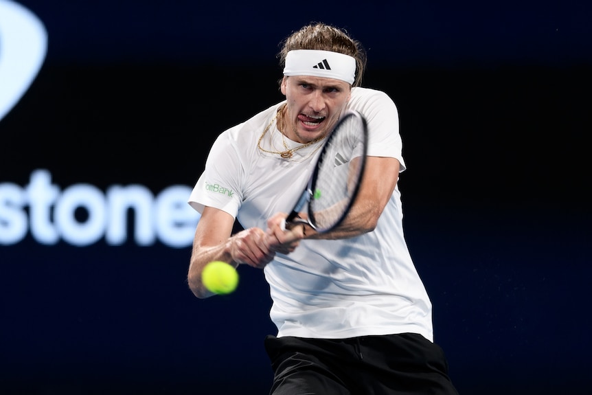 A man wearing a white t-shirt and sweat band hits a tennis ball.