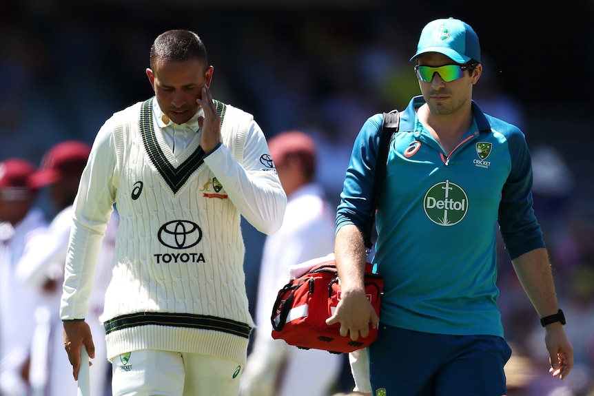 Usman Khawaja looks down and touches the side of his face as he walks off the field with a team doctor
