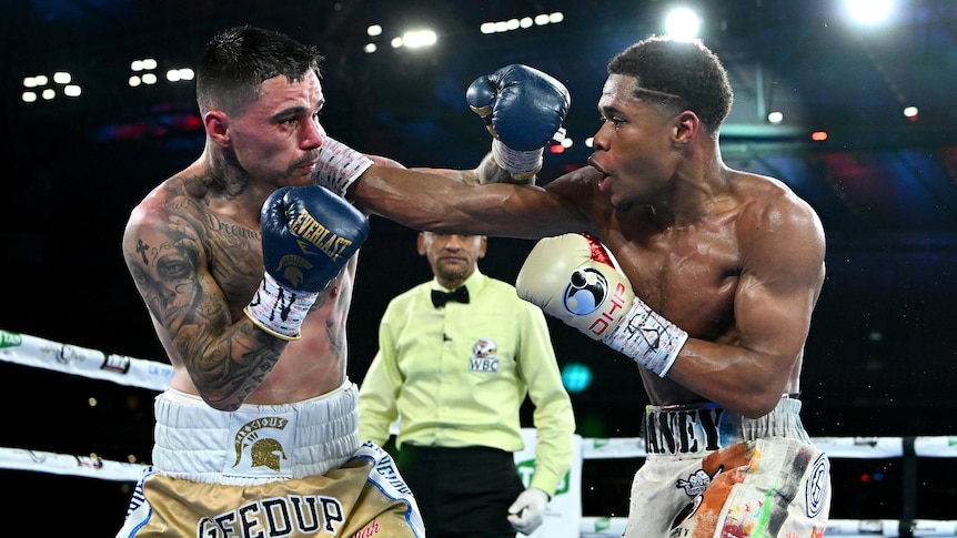 An American boxer reaches out with a right-hand punch against an Australian boxer in the ring, as a referee watches behind them.