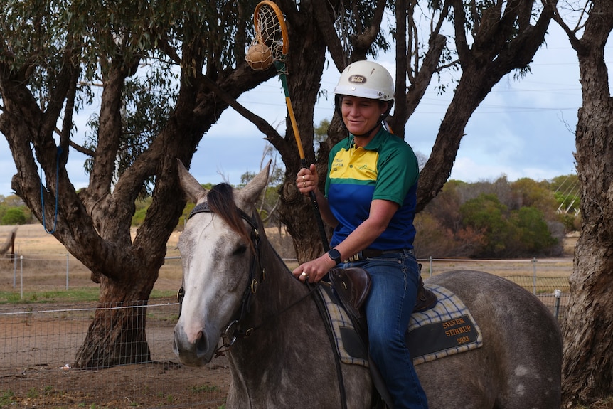 A woman rides a dappled grey horse one-handed, holding a polocrosse stick, outside. She wears blue jeans and a helmet.