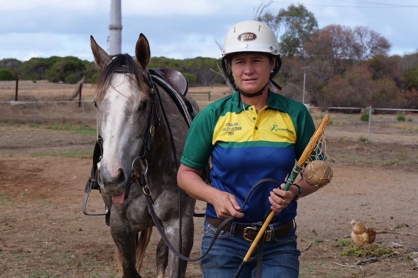 A woman in a riding helmet holds a polocrosse stick and leads a horse with a saddle and bridle, outside on a dusty property. 