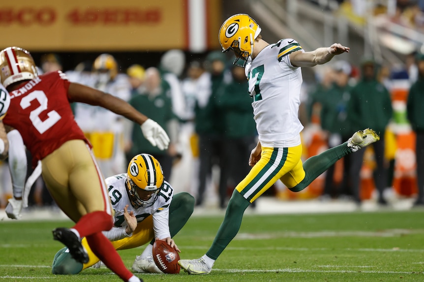 An NFL kicker is about to kick a field goal, a place holder has the ball on the ground, a defender rushes attempt to block