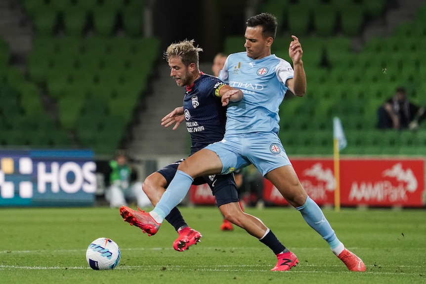 A blonde man wearing navy wrestles another man wearing light blue for a soccer