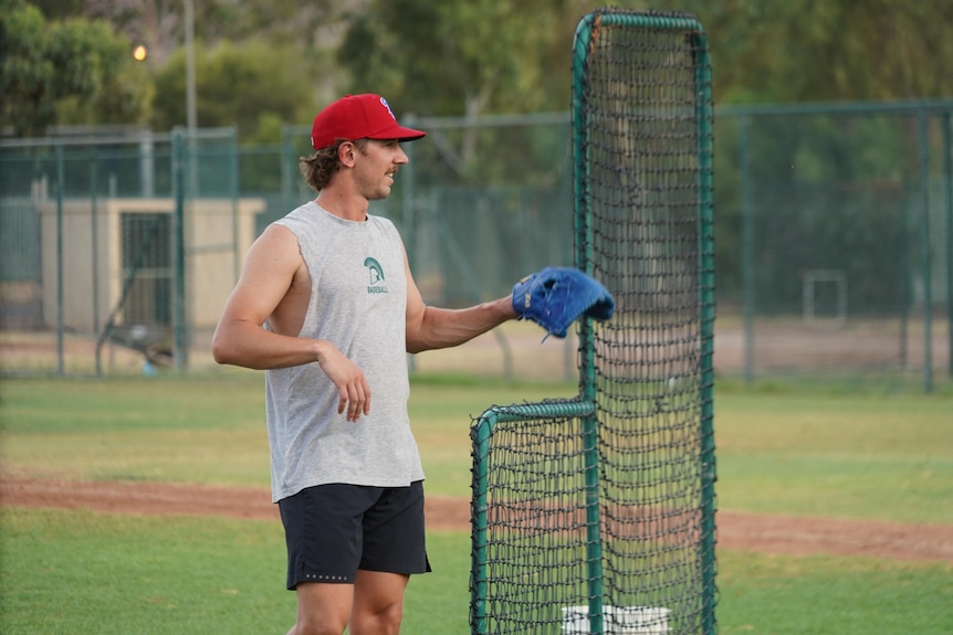 A man wearing a singlet and cap stands in a baseball diamond field with a blue mitten waiting for a ball.