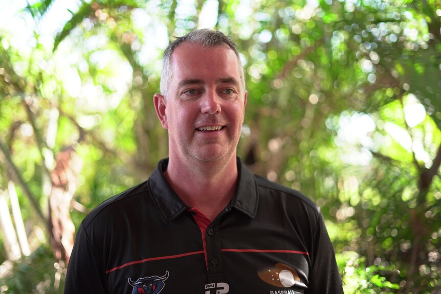 A man in a black polo shirt stands in front of a small bush 