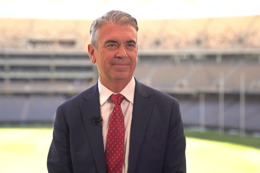 A man in a black suit jacket, white shirt and red tie standing in a sports stadium