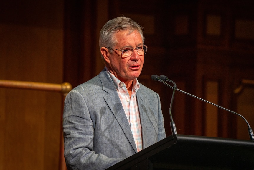 A grieving middle-aged man speaking at a lecturn on stage