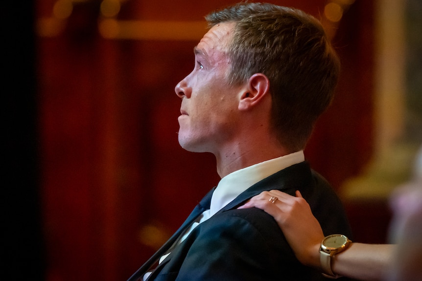 A man upset while looking up. A hand placed on his shoulder in comfort.