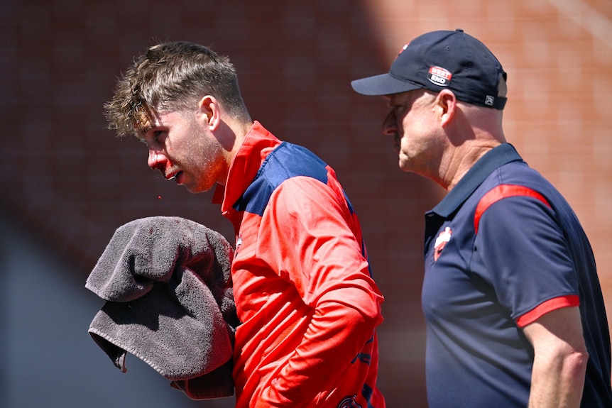 A South Australian cricketer holds a towel under his face as he walks off the ground bleeding, next to a team assistant.