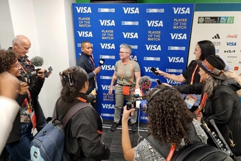 A number of journalists are gathered for a press conference, a person stands in front of a blue backdrop for an interview