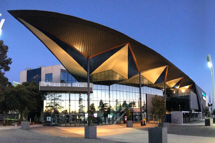 The exterior of a stadium at dusk, with the flood lights on and Geelong Cats branding on the side.