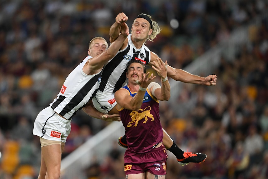 A Collingwood player leaps high and punches the ball clear as he is one of three players grimacing in a contest.