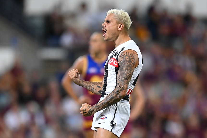 A Collingwood AFL player stands side-on to the camera, shouting in celebration as he pumps his fists.