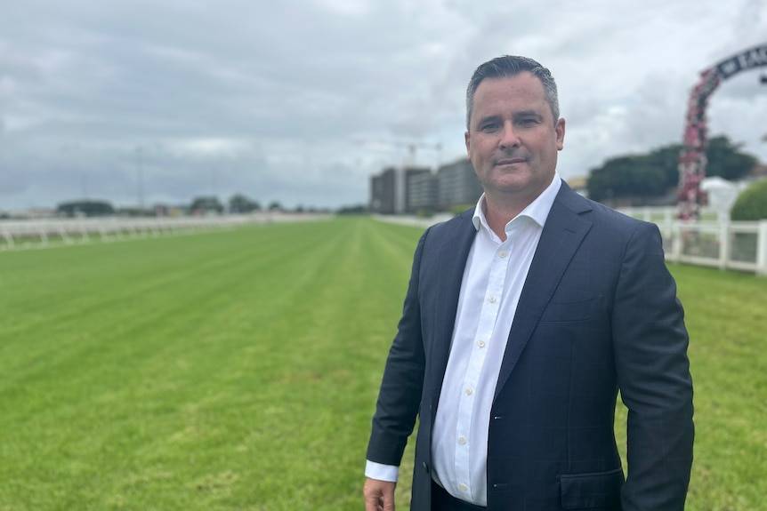 A slightly smiling middle-aged man wearing a white shirt and suit jacket with green grass, high rises in the background.